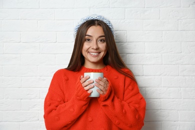 Beautiful young woman in warm sweater with cup of hot drink near white brick wall