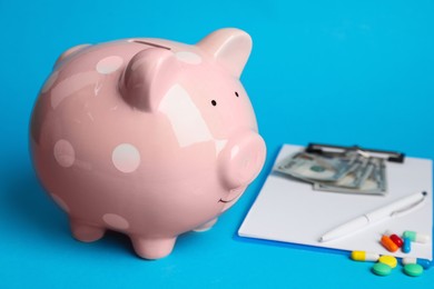 Photo of Pale pink ceramic piggy bank, clipboard and pills on light blue background. Medical insurance