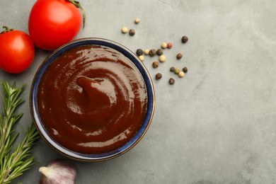 Tasty barbeque sauce in bowl, rosemary, garlic, tomato and peppercorns on grey textured table, flat lay. Space for text