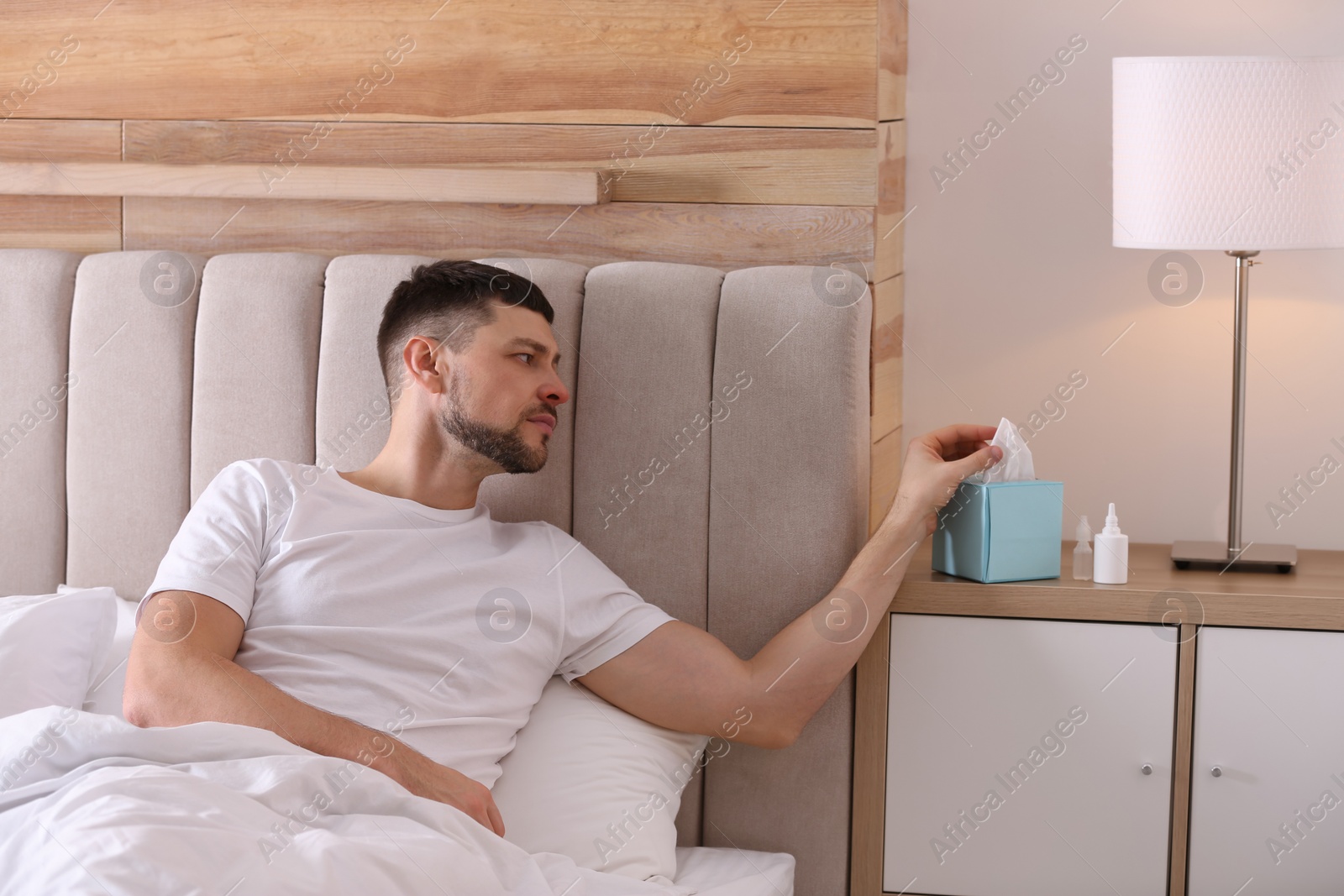 Photo of Ill man with nasal spray, drops and box of paper tissues in bed at home