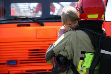 Photo of Firefighter in uniform holding rescued little girl near fire truck outdoors, space for text. Save life