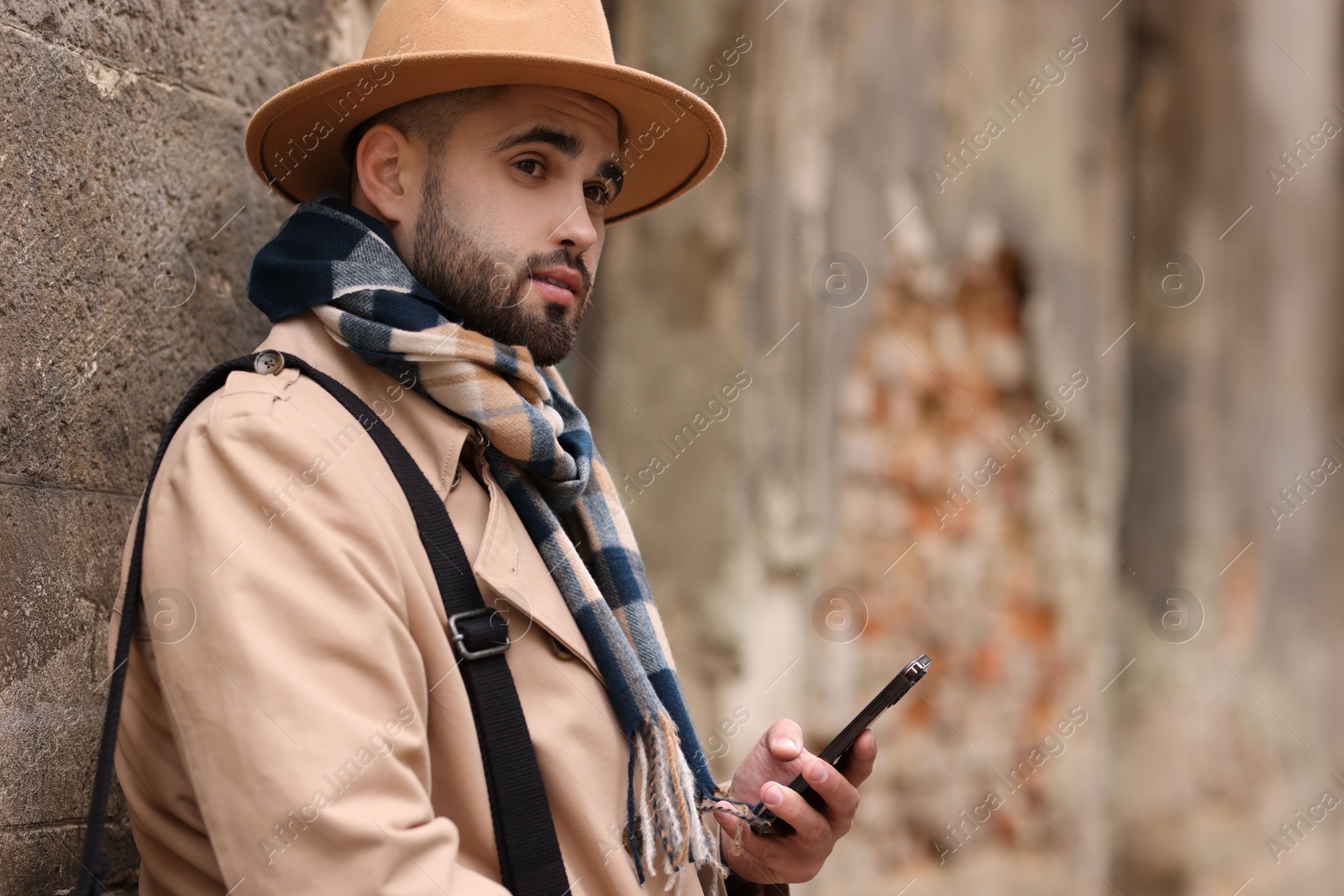 Photo of Handsome man in warm scarf with smartphone near wall outdoors. Space for text