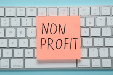 Photo of Paper note with phrase Non Profit and keyboard on light blue background, top view