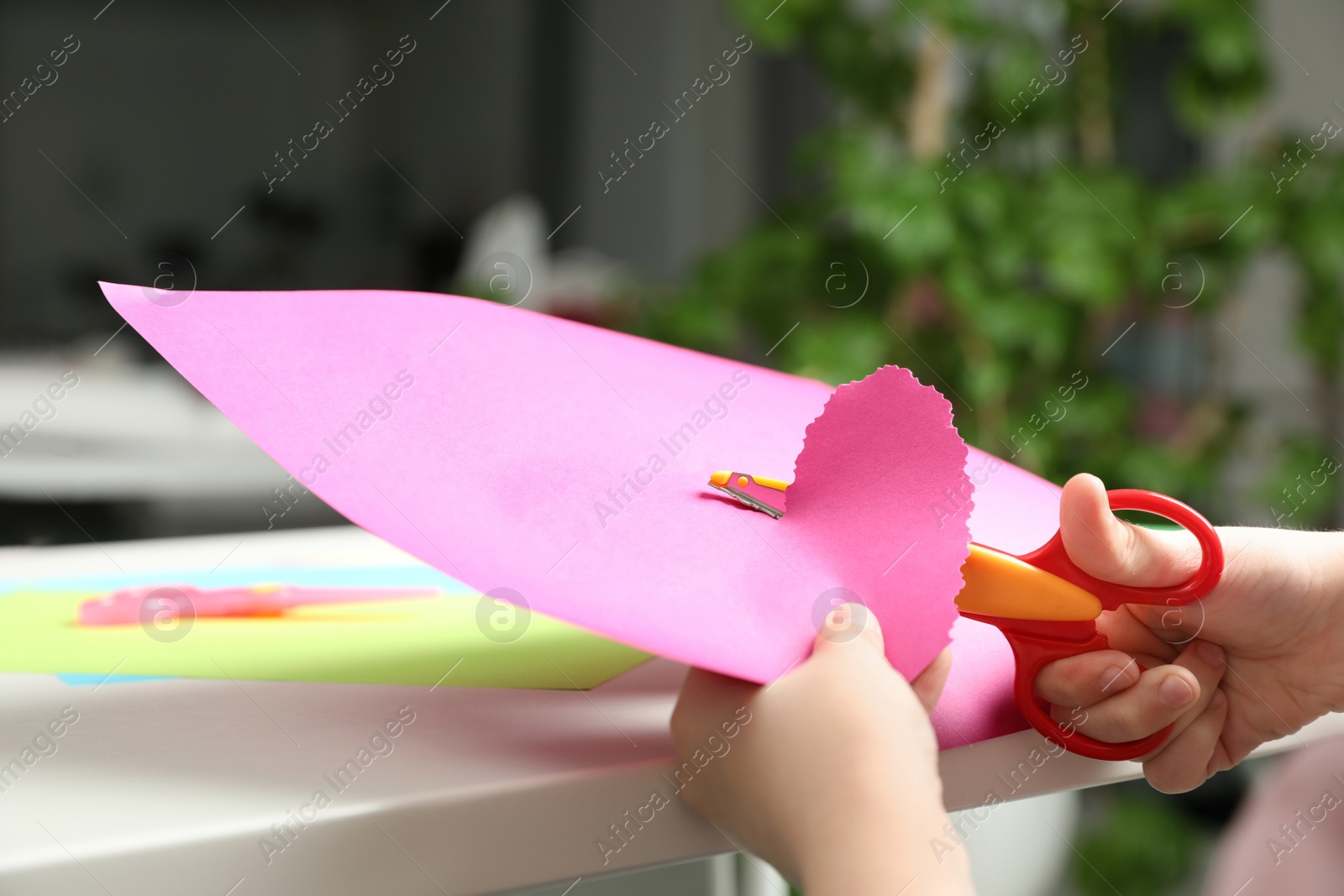 Photo of Child cutting out paper heart with craft scissors at table indoors, closeup