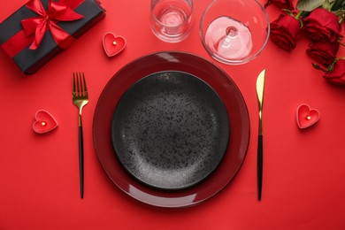 Photo of Place setting with heart shaped candles, gift box and bouquet of roses on red table, flat lay. Romantic dinner