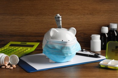 Photo of White ceramic piggy bank with mask, money and pills on wooden table. Medical insurance