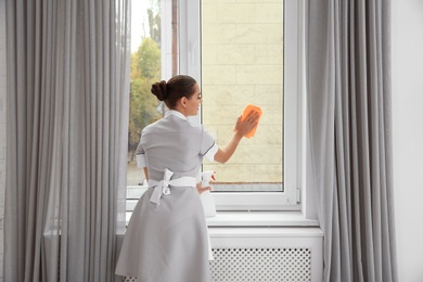 Photo of Young chambermaid cleaning window with rag indoors
