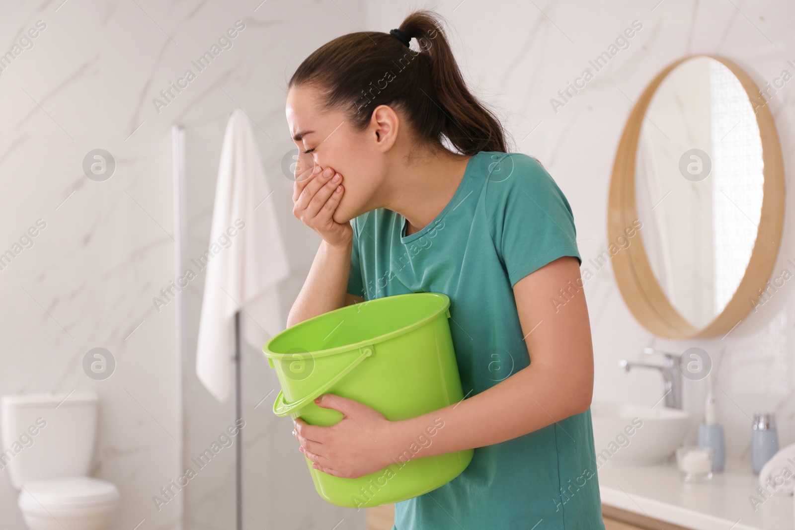 Photo of Young woman with bucket suffering from nausea in bathroom. Food poisoning