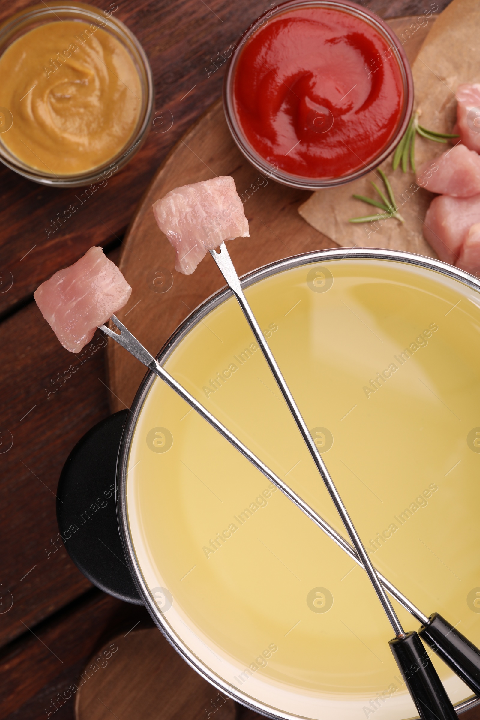 Photo of Fondue pot, forks with pieces of raw meat and sauces on wooden table, flat lay