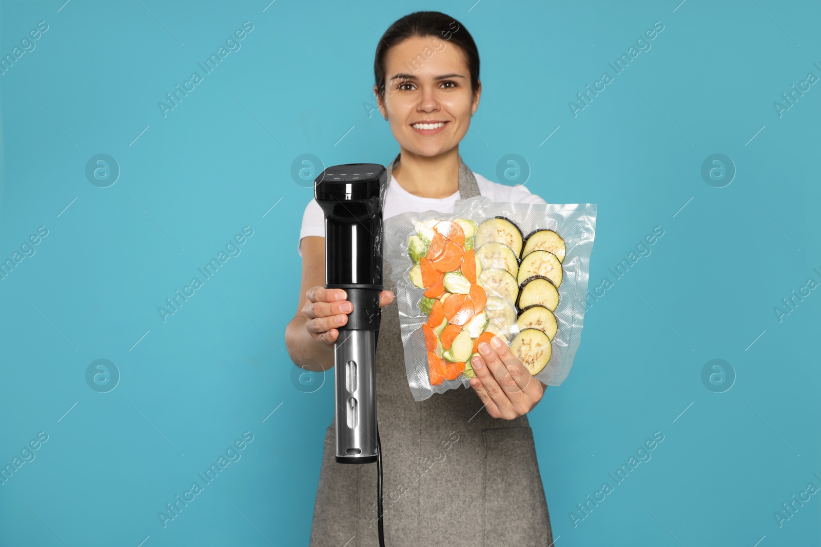 Photo of Beautiful young woman holding sous vide cooker and vegetables in vacuum packs on light blue background.