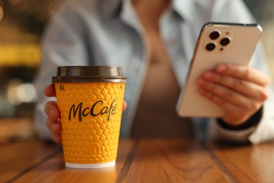 Photo of Lviv, Ukraine - September 26, 2023: Woman with hot McDonald's drink and smartphone at wooden table in cafe, selective focus. Space for text