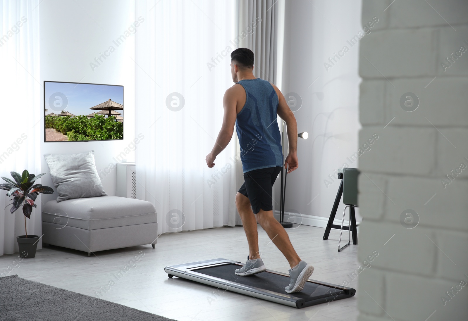 Image of Sporty man training on walking treadmill and watching TV at home