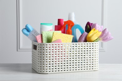 Photo of Spring cleaning. Basket with detergents, flowers and tools on white wooden table