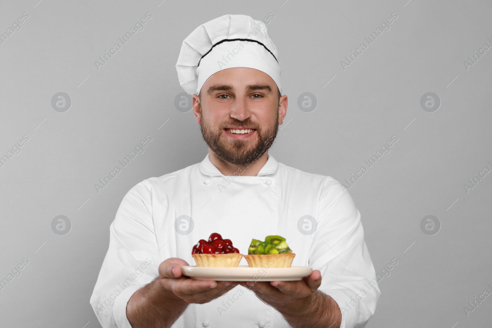 Photo of Happy professional confectioner in uniform holding plate with delicious tartlets on light grey background