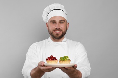 Photo of Happy professional confectioner in uniform holding plate with delicious tartlets on light grey background