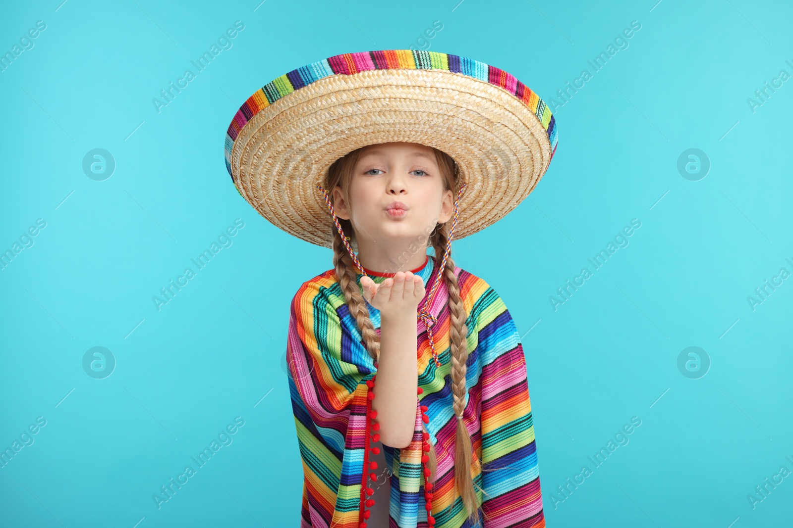 Photo of Cute girl in Mexican sombrero hat and poncho blowing kiss on light blue background