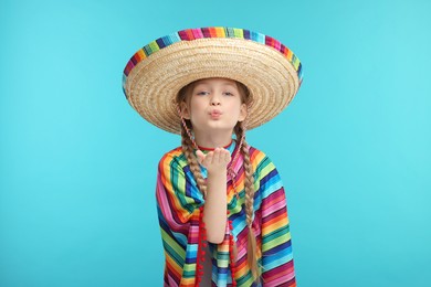 Photo of Cute girl in Mexican sombrero hat and poncho blowing kiss on light blue background