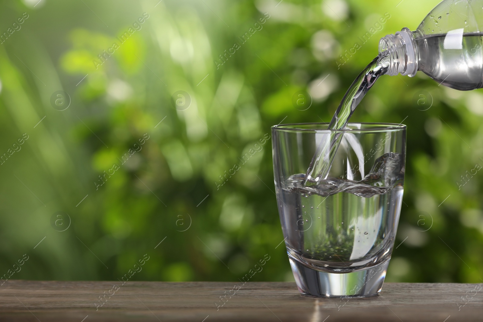 Photo of Pouring water from bottle into glass on wooden table outdoors, closeup. Space for text