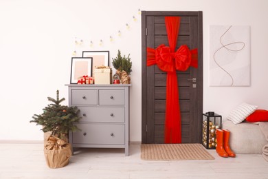 Photo of Beautiful fir tree near chestdrawers and wooden door decorated with red bow in room. Christmas decoration