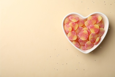 Tasty jelly candies in heart shaped bowl on beige background, top view. Space for text