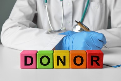 Word Donor made of wooden cubes. Doctor writing in notebook at white table, closeup