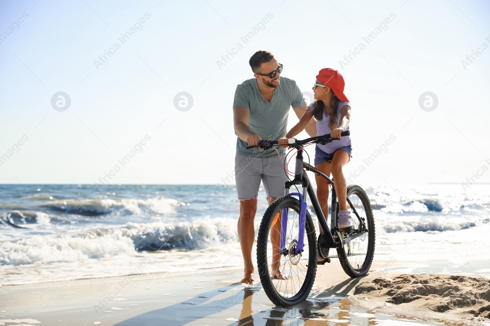 Photo of Happy father teaching daughter to ride bicycle on sandy beach near sea