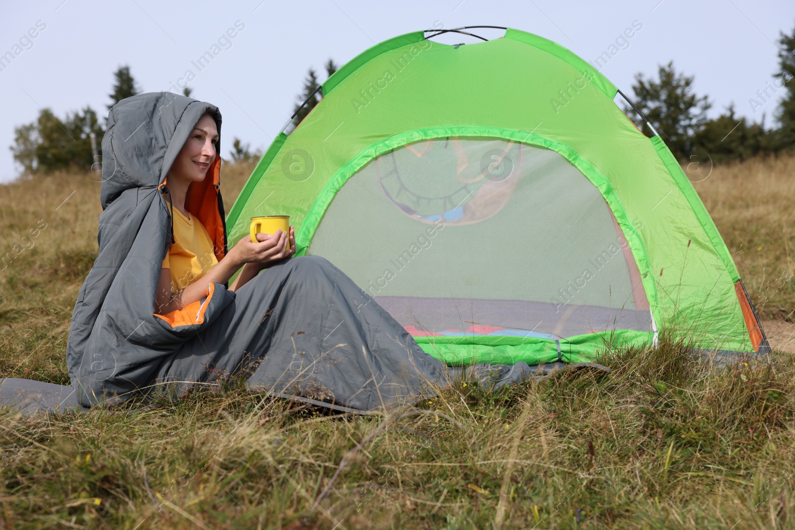 Photo of Mature woman with cup of drink in sleeping bag near camping tent on hill