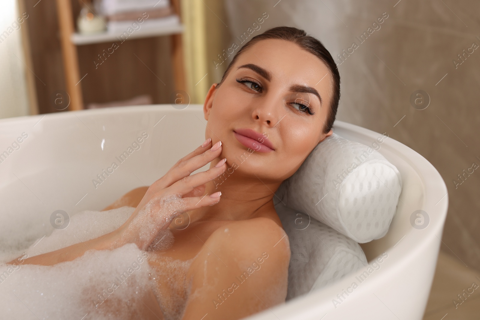 Photo of Young woman using pillow while enjoying bubble bath indoors