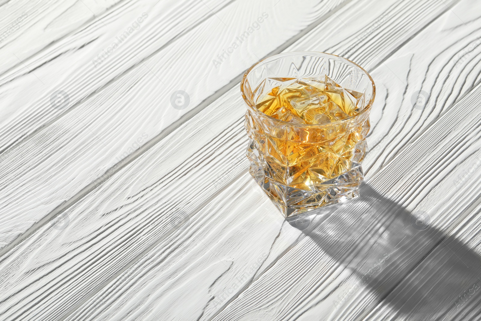 Photo of Whiskey with ice cubes in glass on white wooden table, space for text