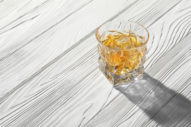 Photo of Whiskey with ice cubes in glass on white wooden table, space for text