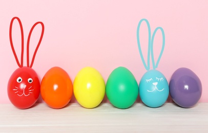 Image of Two eggs with drawn faces and ears as Easter bunnies among others on white wooden table against pink background