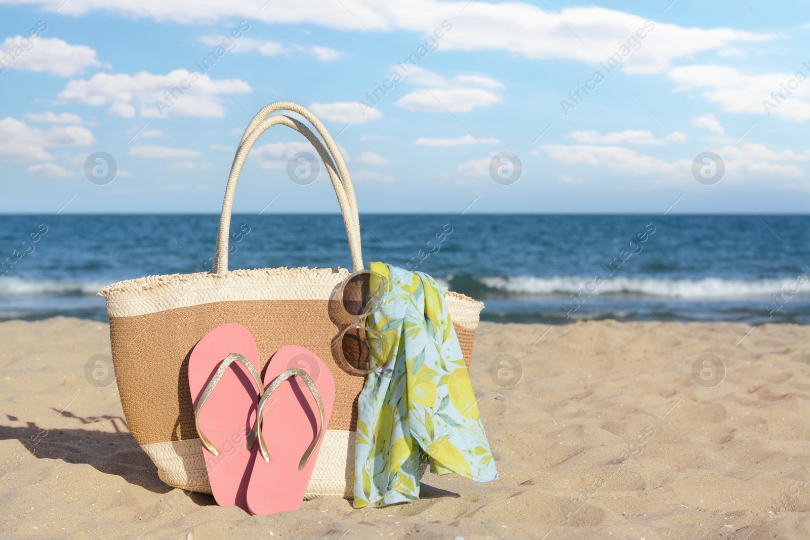 Photo of Straw bag with beach wrap, sunglasses and flip flops on sandy seashore, space for text. Summer accessories