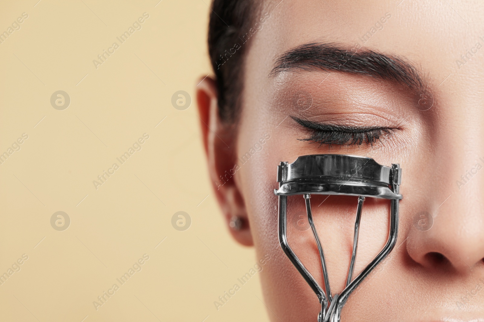 Photo of Young woman using eyelash curler on beige background, closeup. Space for text