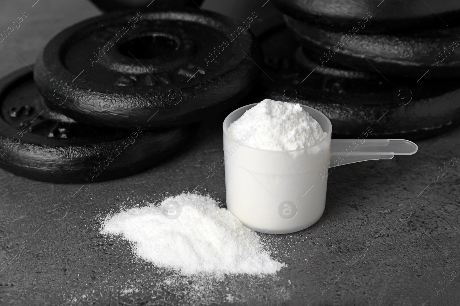 Photo of Protein powder, measuring scoop and dumbbell plates on grey table. Space for text