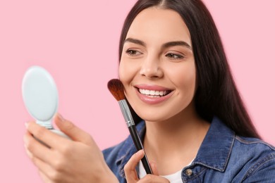 Beautiful woman with cosmetic pocket mirror applying makeup on pink background