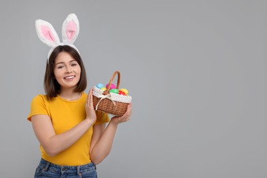 Photo of Easter celebration. Happy woman with bunny ears and wicker basket full of painted eggs on grey background, space for text