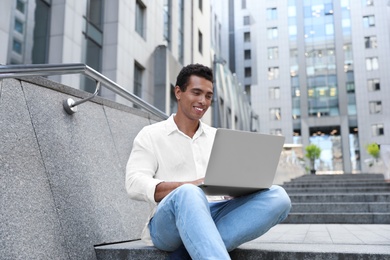 Handsome young African-American man with laptop sitting on stairs outdoors