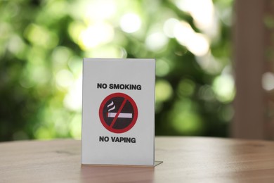 Photo of No Smoking No Vaping sign on wooden table against blurred background