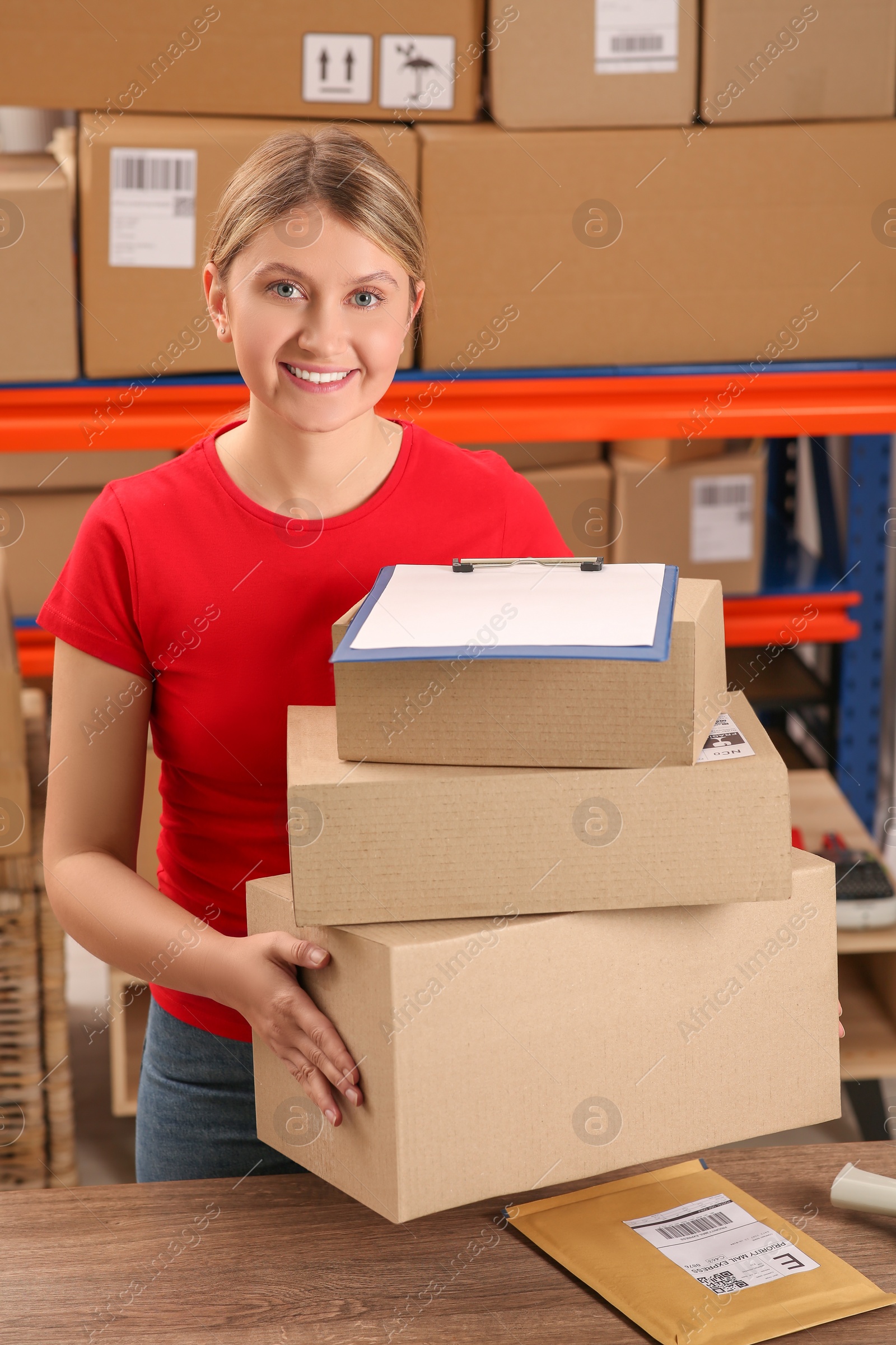 Photo of Post office worker with parcels and clipboard near rack indoors