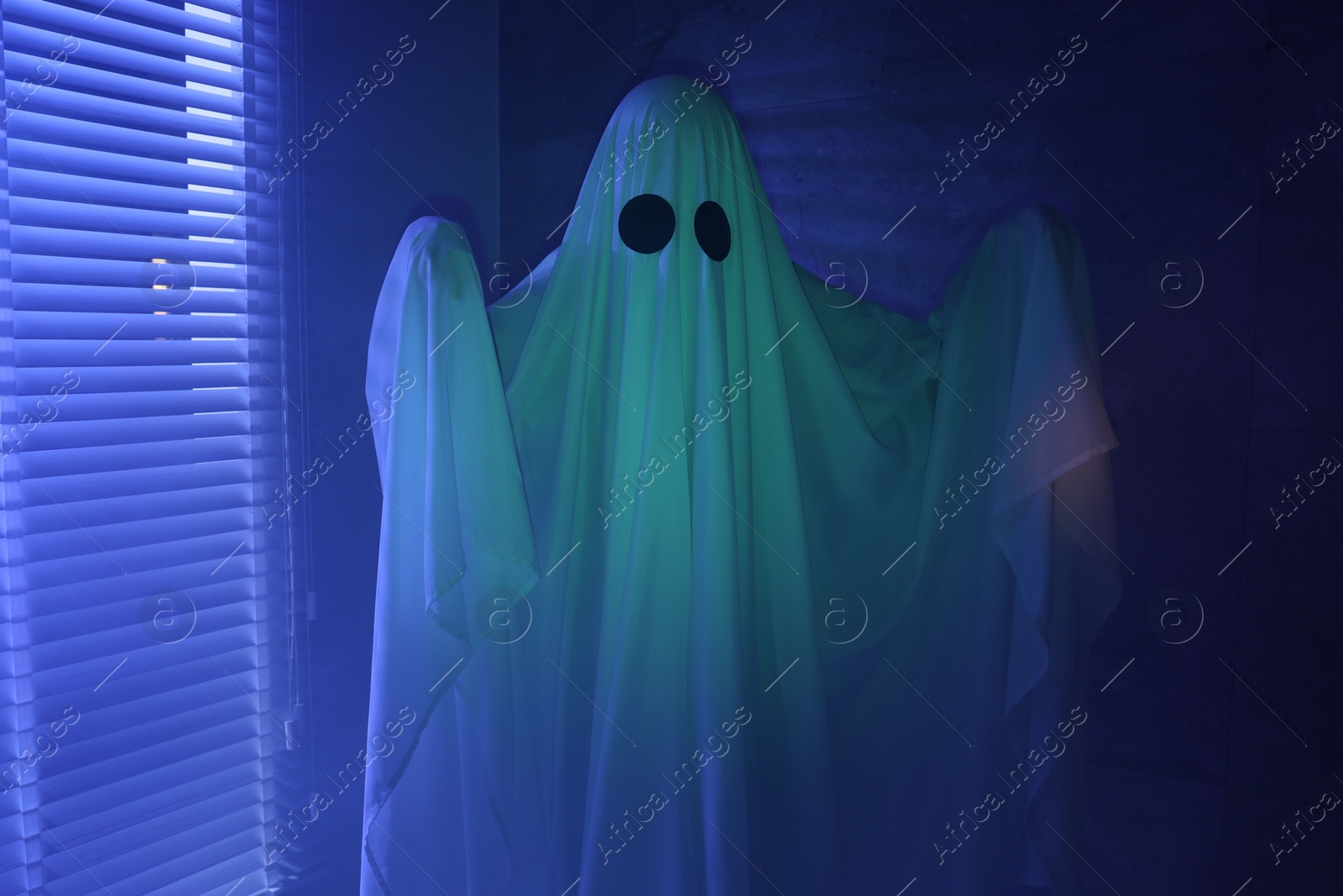 Photo of Creepy ghost. Woman covered with sheet near window in blue light