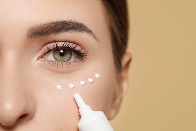 Young woman applying cream under eyes on beige background, closeup. Space for text