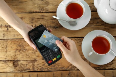 Woman with credit card using modern payment terminal at wooden table, top view