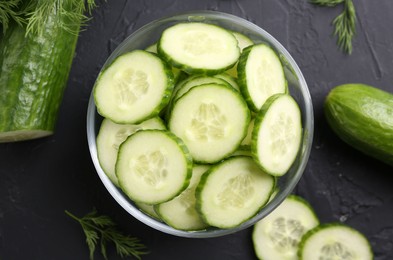 Photo of Cut cucumber in glass bowl, fresh vegetables and dill on dark gray textured table, flat lay