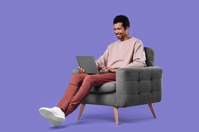 Happy man with laptop sitting in armchair on purple background
