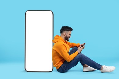 Image of Man with mobile phone sitting near huge device with empty screen on light blue background. Mockup for design