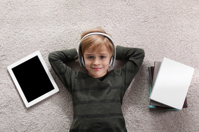 Cute little boy with headphones and tablet listening to audiobook on floor indoors, flat lay