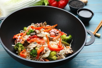 Stir fried noodles with mushrooms, seafood and vegetables in wok on light blue wooden table, closeup