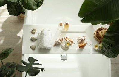 Bath tray with spa products, towels and shells on tub in bathroom, top view