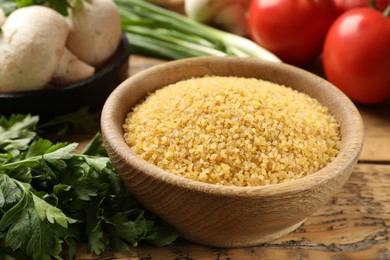 Photo of Raw bulgur in bowl, parsley and vegetables on wooden table, closeup