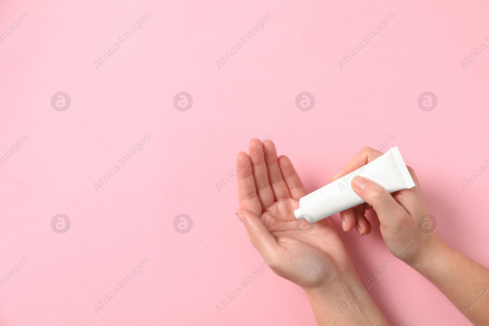 Photo of Woman applying cosmetic cream from tube onto her hand on pink background, top view. Space for text